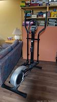 Elliptical machine in a room, showing the side view with the flywheel and handle bars.