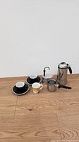 Photo showing three espresso cups and saucers in black and white and yellow design, along with a small stainless steel milk frothing pitcher, a stainless steel stovetop espresso maker, a manual espresso coffee maker with brass spout, and a black handled tool.