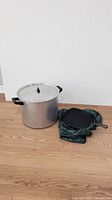 Photo showing a large aluminum pot with lid and black handles next to a cast iron pan with a handle, partially covered by a green padded protective cover, placed on a wooden floor against a white wall.