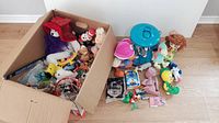 Photo showing two groups of toys: one inside an open cardboard box with plush and other toys, other group outside including a blue plastic jail/playset and various small toys.