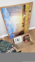 Box containing several framed and unframed picture items next to a large Grand Canyon Nature poster leaning on the wall.