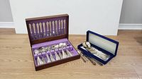 Full view of two sets of silver cutlery with cases, one wooden-lined purple, one blue velvet-lined.