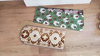 Two table runners shown on wooden floor: one green with daisies and ladybugs, one brown and beige geometric with rounded edges.