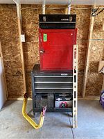 View of stacked tool boxes including a red cabinet, large Stanley tool chest, a black Alltrade toolbox, with a white spirit level and yellow metal stand propped up