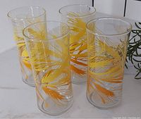 Four clear glass tumblers decorated with a yellow and orange wheat pattern, placed on a white surface with some greenery in the background.