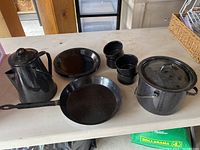 Full view of all items on white table: 4 plates, 4 mugs, small cooking pot with lid, coffee pot, and fry pan with bent handle.