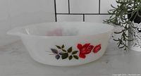 Side angled view of the white opaque Pyrex casserole dish with June Rose rose floral pattern, placed on a white countertop with a plant in the background.