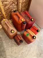 Photo of six used plastic gas cans on concrete floor against plywood wall. Three large and three smaller cans in red and orange colors with yellow and black caps.