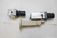 Two Samsung surveillance cameras displayed on a white surface showing one camera laid flat with black lens mounted on a beige mounting bracket and the other without bracket.