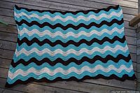 Full view of the large handmade crochet blanket laid flat on wooden deck, showcasing wavy stripes in black, blue, and white.