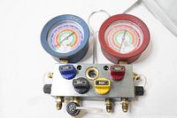 View of the entire aluminum manifold gauge set showing two large pressure gauges with blue and red protective casings, four control valves with color-coded handles, and brass hose fittings.
