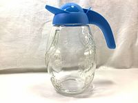 Front view of the vintage glass pitcher with blue lid and handle showing embossed fruit patterns on clear glass.