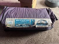 Purple roller suitcase bag containing the tent, showing the worn and torn label with brand, series name, and tent capacity details visible.