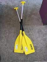 Photo shows two yellow Propel kayak paddles with silver shafts crossed on carpeted floor.