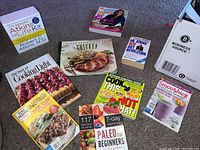 Photo showing 10 healthy cooking books displayed on carpet including Atkins diet, paleo beginners, smoothie recipe cards, and holiday recipes.