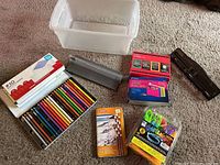 Top view showing colored pencils, pastel pencils, craft laces, envelopes, hole punch, and storage box arranged on carpet.