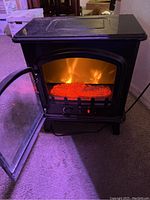 Photo showing the front of the black electric heater stove with glass door open, glowing red faux logs and flame effect visible inside.