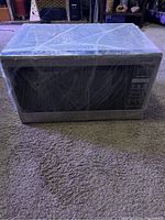 Front view of Panasonic metal silver microwave oven wrapped in plastic on carpeted floor.
