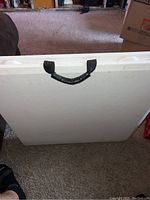 Folded white plastic table showing black carrying handle attached with black strap.