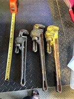 Three large pipe wrenches with a measuring tape showing lengths around 18-22 inches laid on a textured metal surface.