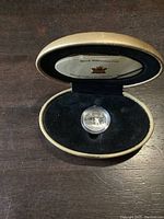Silver coin in protective capsule displayed inside a beige oval case with a navy blue velvet interior.