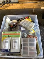 Top view of blue container showing assortment of maintenance supplies including packaged light switches, compartment organizer, tape rolls, and cable clips.