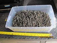 Clear plastic container filled about 3/4 with metal screws. Yellow measuring tape in the view for scale shows about 13 inches length of the container.