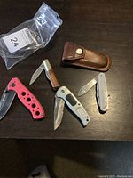 Overview of five pocket knives arranged around one brown leather case on dark wood surface.