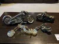 Four motorcycle collectibles displayed on a dark table including glass, metal, and a clock motorcycle.