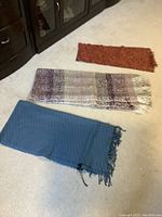 Photo showing the three women's wraps laid out flat on a carpet floor: rust-colored with fringe, multi-colored in browns and cream with fringe, and solid blue with fringe.