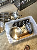 View of clear plastic bin containing small stainless steel pots, handheld grater, muffin tray, wooden rolling pin with handles