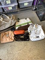 Photo showing seven assorted purses and bags laid out on a metal textured floor, including gray handbag, white handbag, tan satchel, black textured bag, striped tote, small orange pouch, and black clutch.