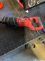 Red Hyper Tough reciprocating saw lying on a black textured workbench surface. The power cord is wrapped around the tool.