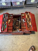 Top view of red metal toolbox opened showing all compartments with tools including chisels, clamps, saw, scissors.