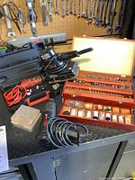 Photo showing Milwaukee rotary tool with red power cord, a black case, wooden boxes of rotary bits and various accessories, electric sander, and heat tool on a black metal workbench.