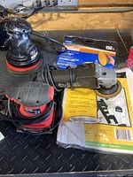 Shows two orbital sanders, angle grinder with wire brush wheel, box of sandpaper, and a part of the coverall packaging.