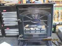 Front view of small black electrical fireplace heater showing glass front with artificial logs inside.