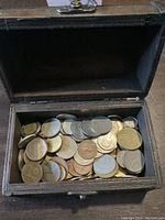 Open wooden chest filled with mixed foreign coins of various sizes and colors.