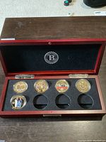 Wooden presentation box open showing 5 gold plated coins in protective capsules set in black foam insert. The box's inside lid has a black velvet lining with a 'B' emblem.