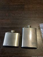 Two stainless steel whiskey flasks on wooden surface, one smaller smooth finish and one larger size
