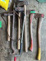 Overview of axes, hammers, and metal spike tool arranged on wooden surface showing wear and paint marks on handles.