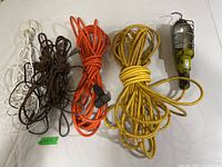 Five extension cords in white, brown, orange, and yellow colors next to a yellow work light with a protective cage and hook, arranged on a white surface.