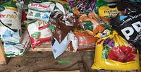 Photo showing a large assortment of fertilizer and soil bags including Scotts Turf Builder and Miracle-Gro potting soil brands on wooden floor.