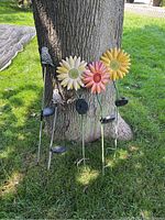 Five garden solar picks leaning against a tree on grass. Three flower designs and two owl figures on metal stakes visible with solar panels at the bottom.