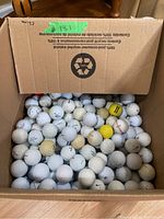 Cardboard box filled with numerous mostly white golf balls with a few yellow balls visible on top.