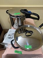 Fresco stainless steel pressure cooker with black handle and lid, accompanied by its user manual and a 10.5" diameter Tramontina frying pan with lid.