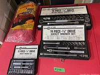 Overview of three socket wrench sets in their cases and a pack of yellow Sundance shop towels on a red surface