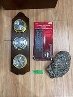 Full lot photo showing Taylor wooden mounted weather station with three brass dials, Taylor packaged indoor/outdoor thermometer, and garden rock on a wooden floor.