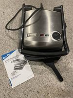 Front view of the closed Breville Gourmet Health Grill showing the stainless steel top, control knob and indicator lights along with instruction manual.