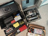 Opened black metal toolbox showing several compartments filled with hand tools such as pliers, screwdrivers, measuring tape, and assorted small hardware.